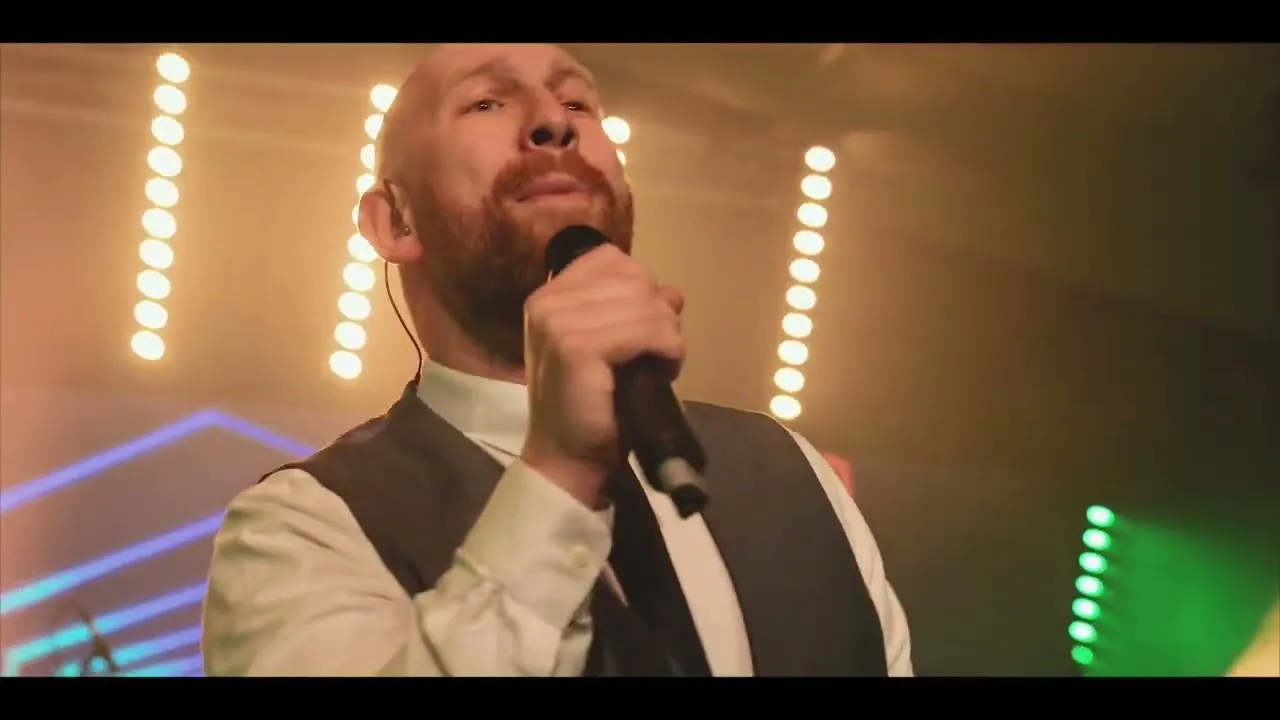 Man with a beard singing into a microphone on stage with bright stage lights in the background.