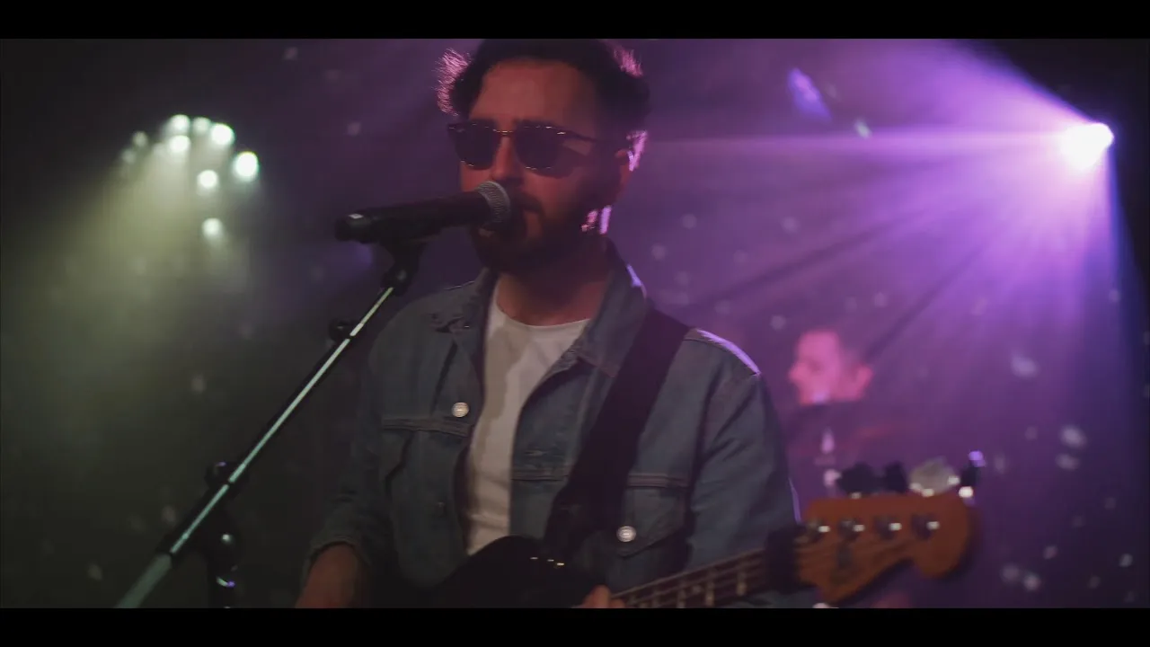 Man wearing sunglasses and denim jacket singing into a microphone while playing electric bass guitar on stage with purple and white stage lights.