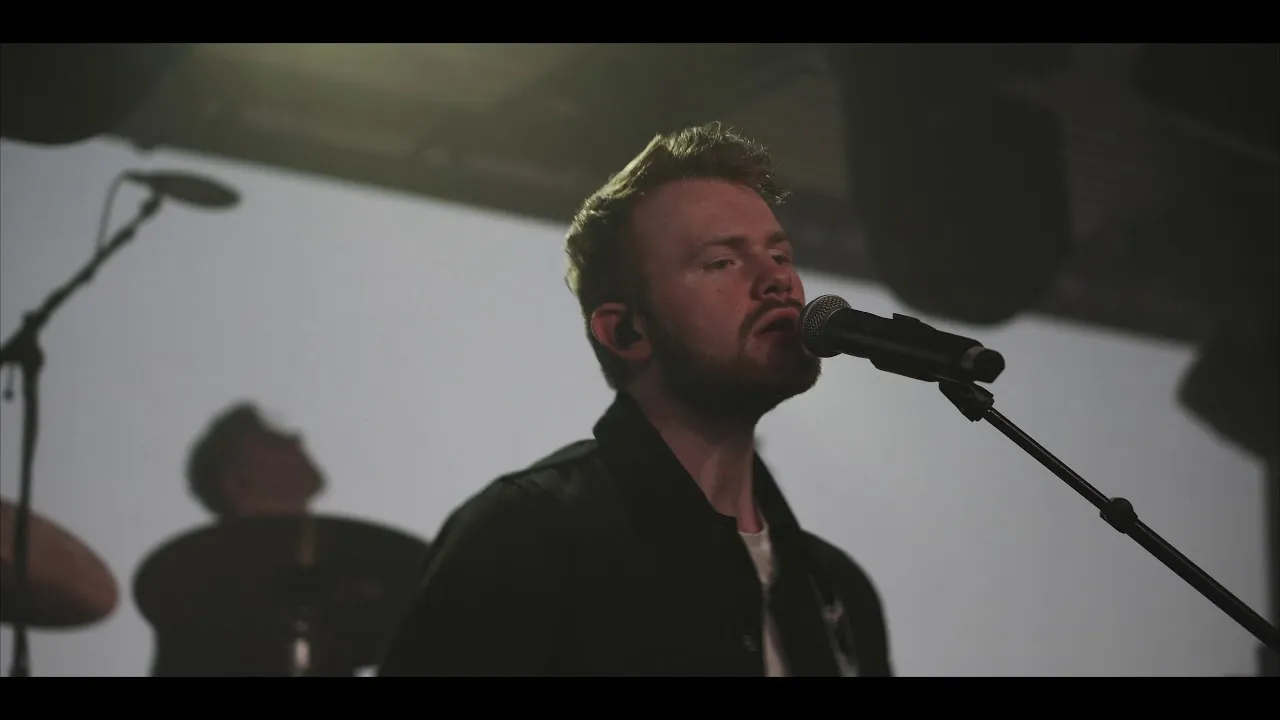 Bearded male singer performing into a microphone with a drummer blurred in the background.