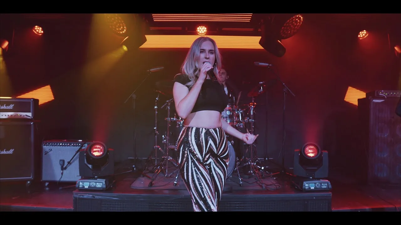 Female singer performing on stage with microphone, wearing a black crop top and patterned skirt, with drummer and stage lights in the background.