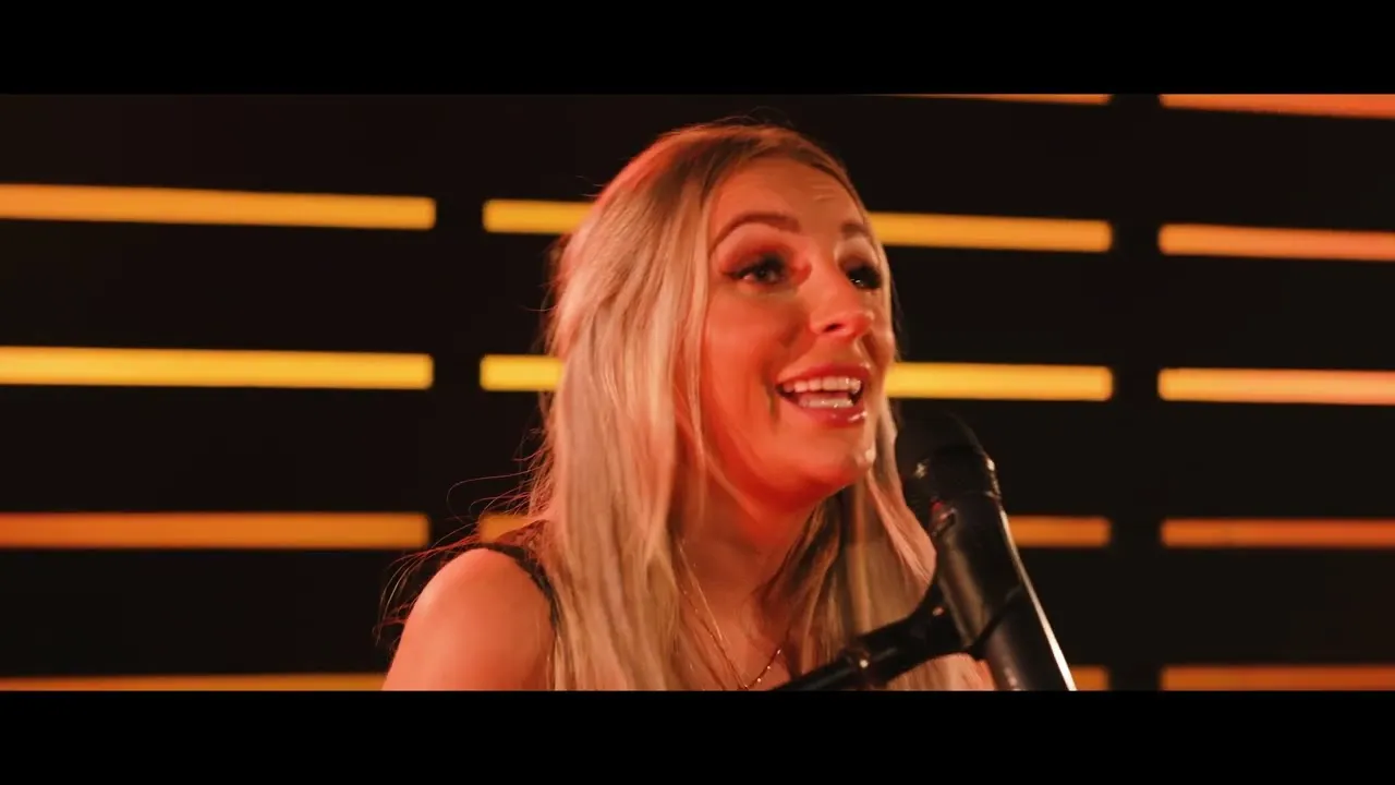 Blonde woman singing into a microphone with warm orange backlight panels behind her.