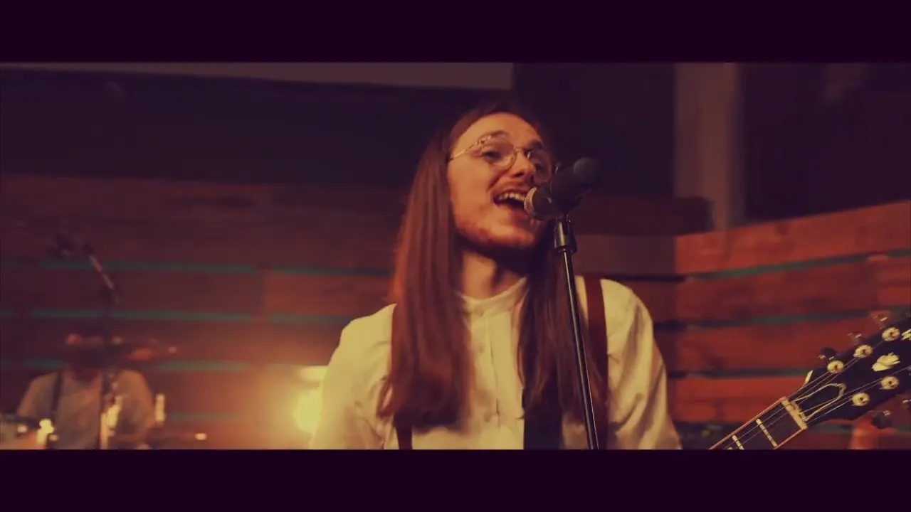Long-haired musician wearing glasses sings into a microphone while playing guitar in a dimly lit wooden room.