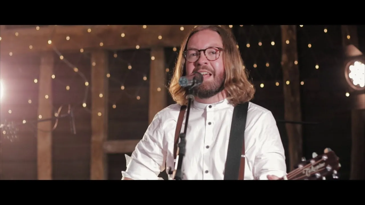 Man with shoulder-length hair and glasses singing into a microphone while playing guitar in a warmly lit room with string lights.
