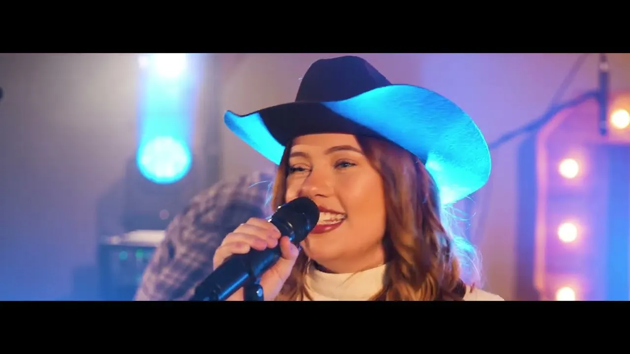 Smiling woman wearing a cowboy hat singing into a microphone with colorful stage lights in the background.