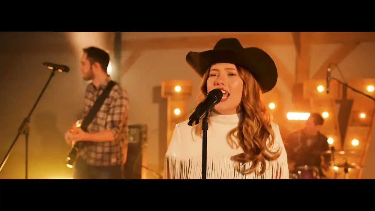 Woman wearing a black cowboy hat singing into a microphone with band members playing guitar and drums in the background.
