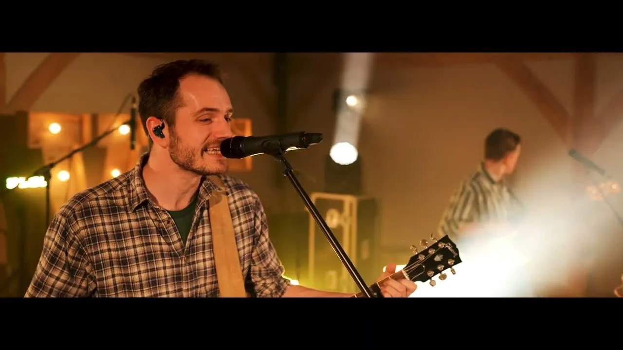 Male musician wearing a plaid shirt singing into a microphone and playing guitar on a warmly lit stage.