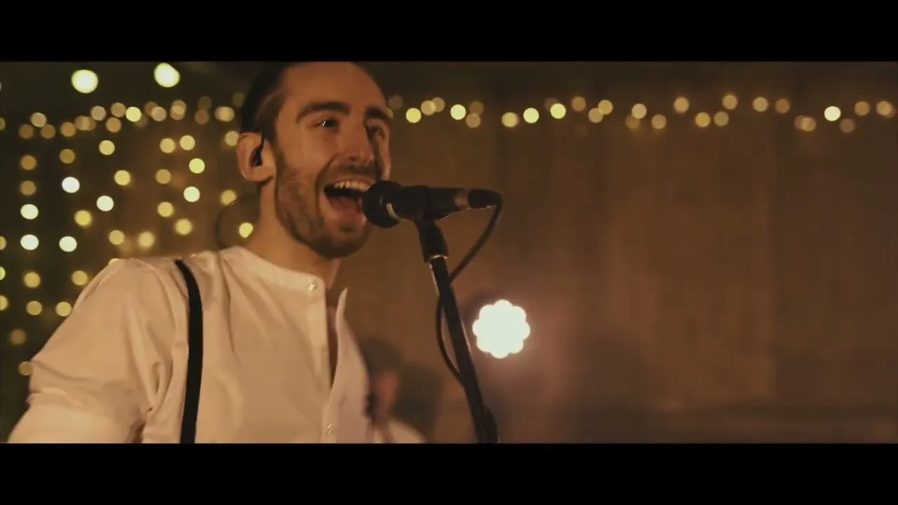 Male singer wearing a white shirt with suspenders, singing into a microphone with warm string lights in the blurred background.