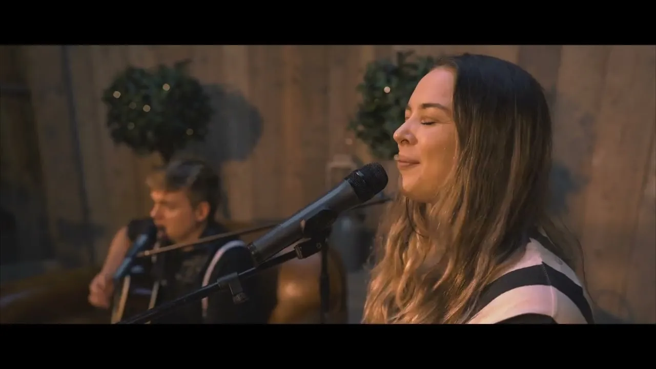 Woman singing into a microphone with eyes closed, man playing guitar in the background.