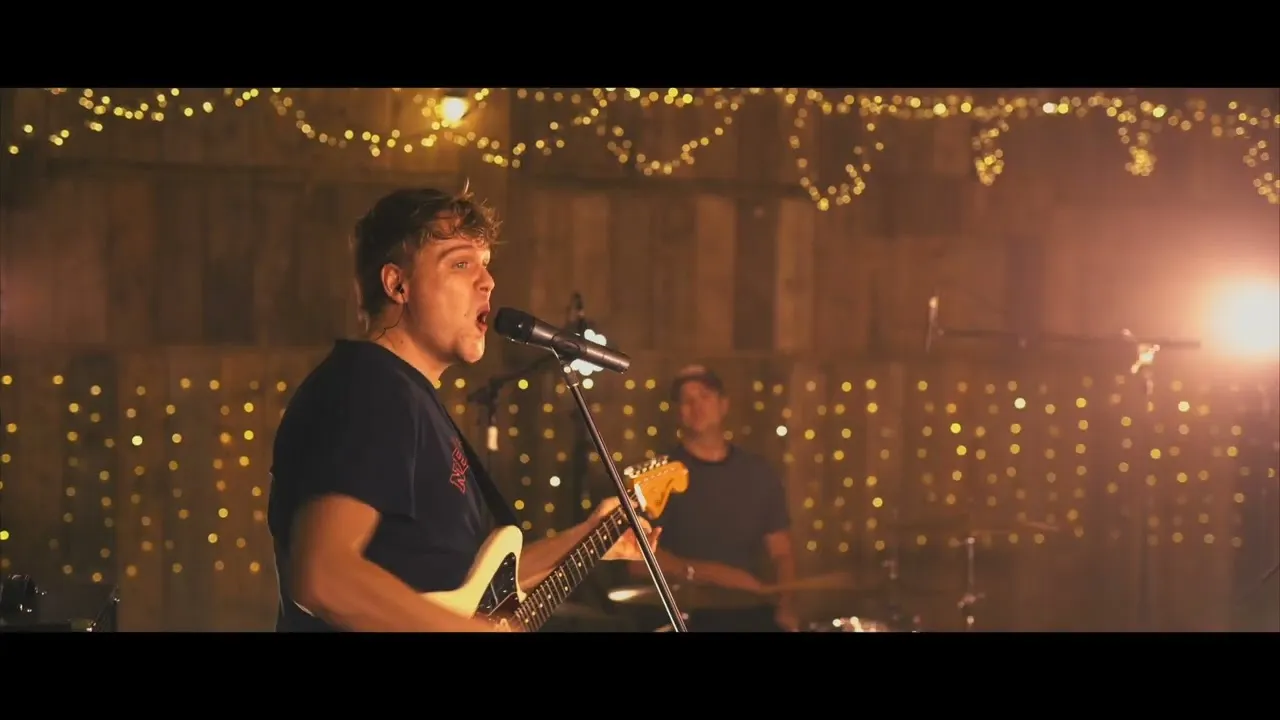 Male musician singing into a microphone and playing electric guitar with drummer in the background and warm string lights hanging behind.