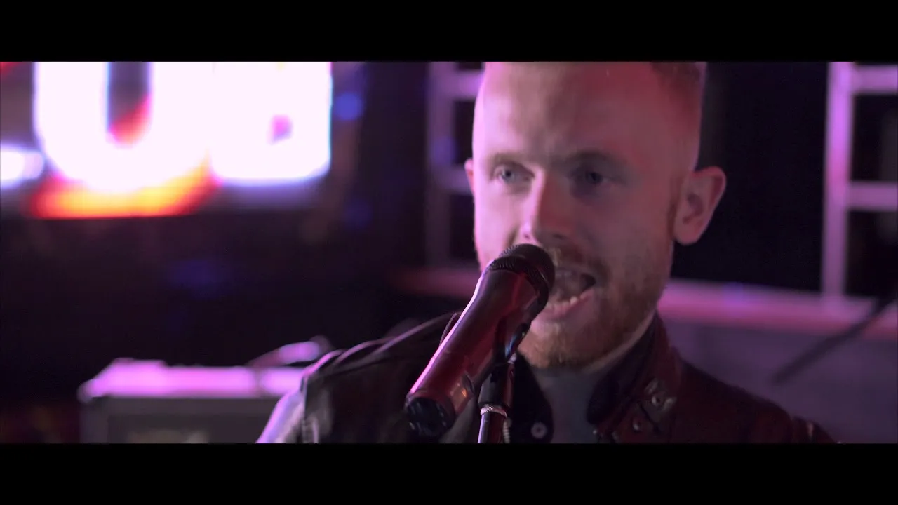 Male singer with short red hair and beard singing into a microphone with a purple and dark background.