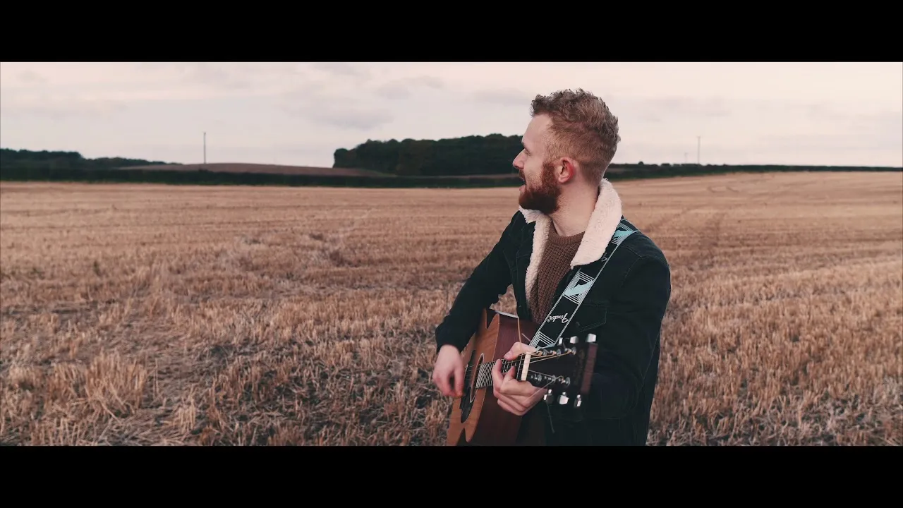 Man with beard playing acoustic guitar in an open field during overcast weather.