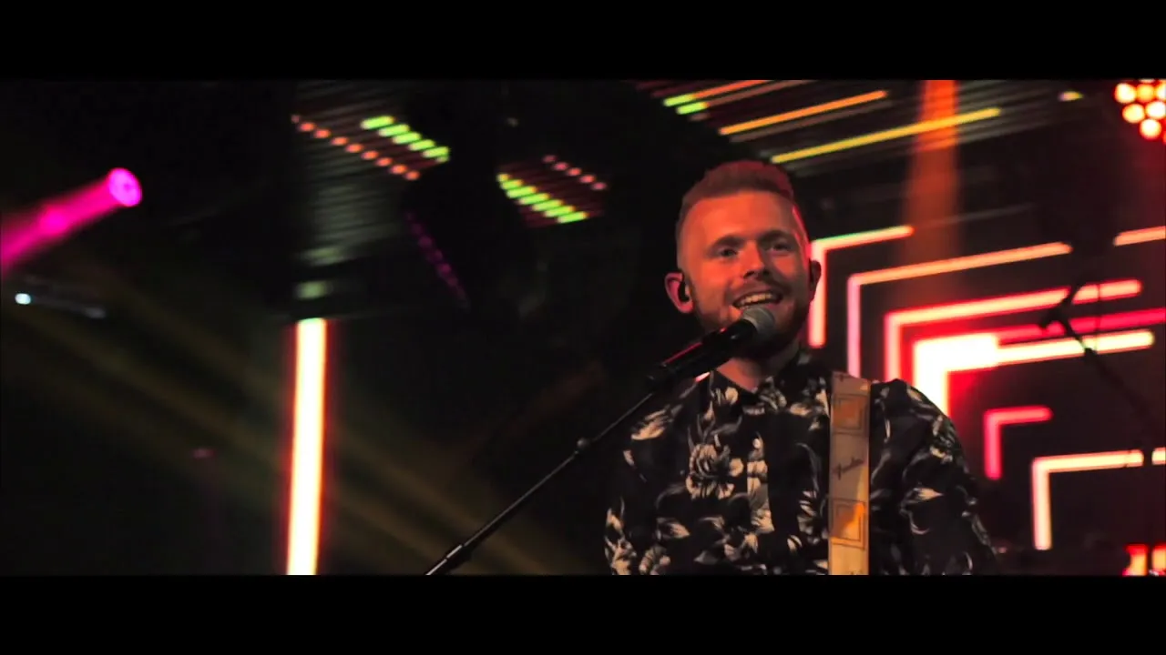 Smiling male musician singing into a microphone on stage with colorful neon lights in the background.