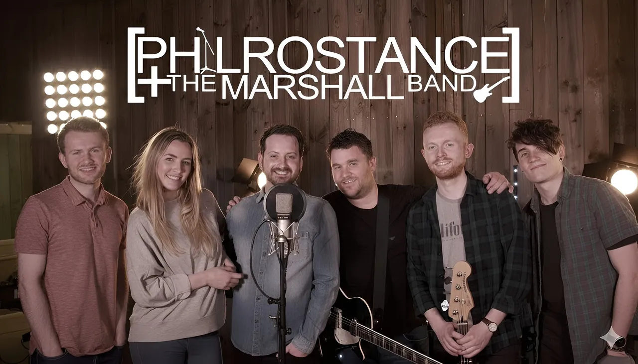 Six members of The Marshall Band standing together in a studio with a microphone and guitar, smiling.