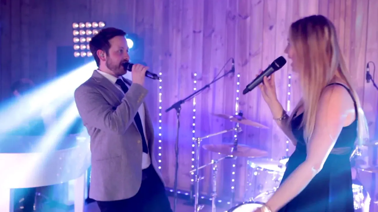 A man in a suit and a woman in a black dress singing into microphones on a stage with purple lighting and musical instruments in the background.