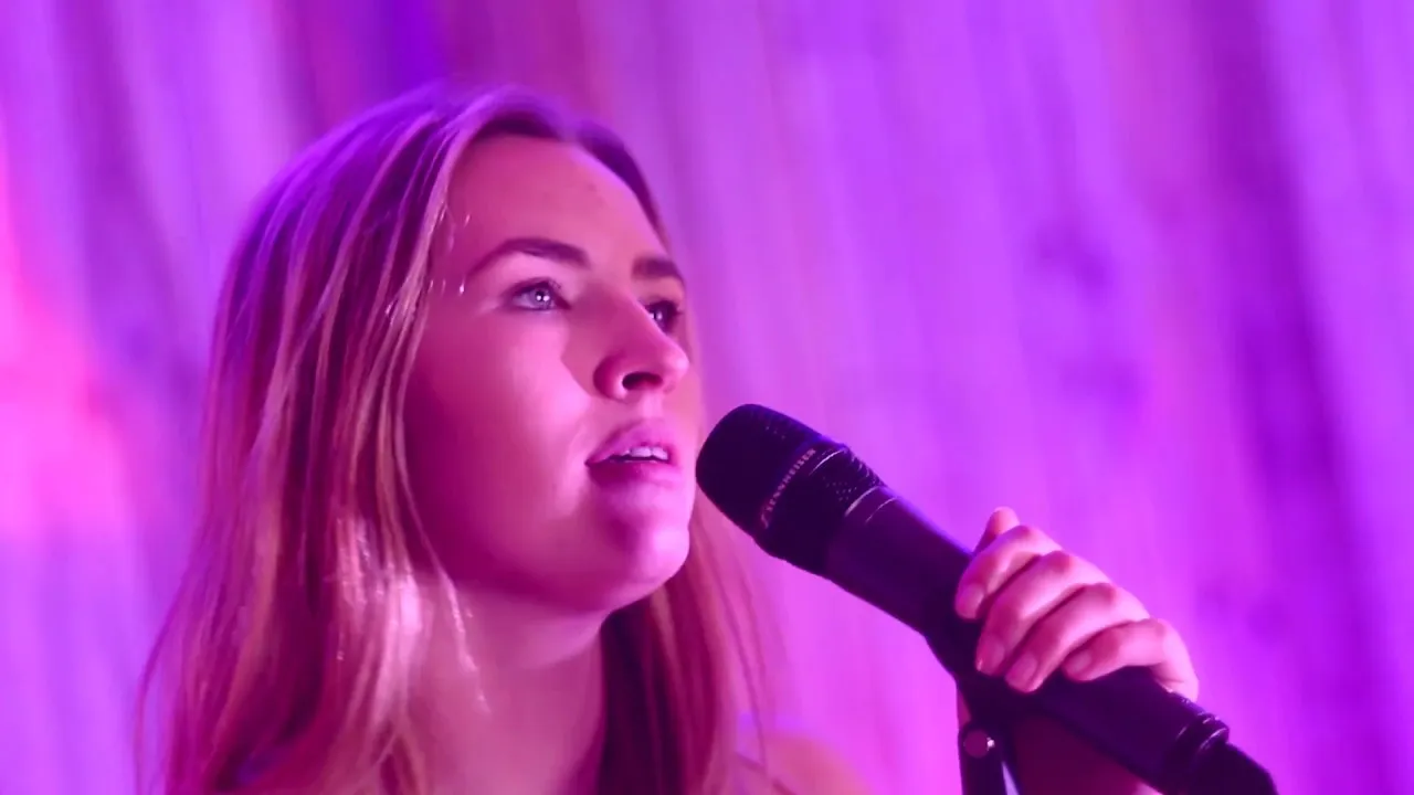Young woman with long blonde hair singing into a microphone against a purple-lit background.