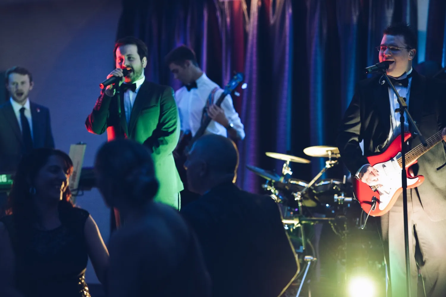 Band performing on stage with two vocalists, a guitarist, a bassist, and a drummer, while people dance in front.