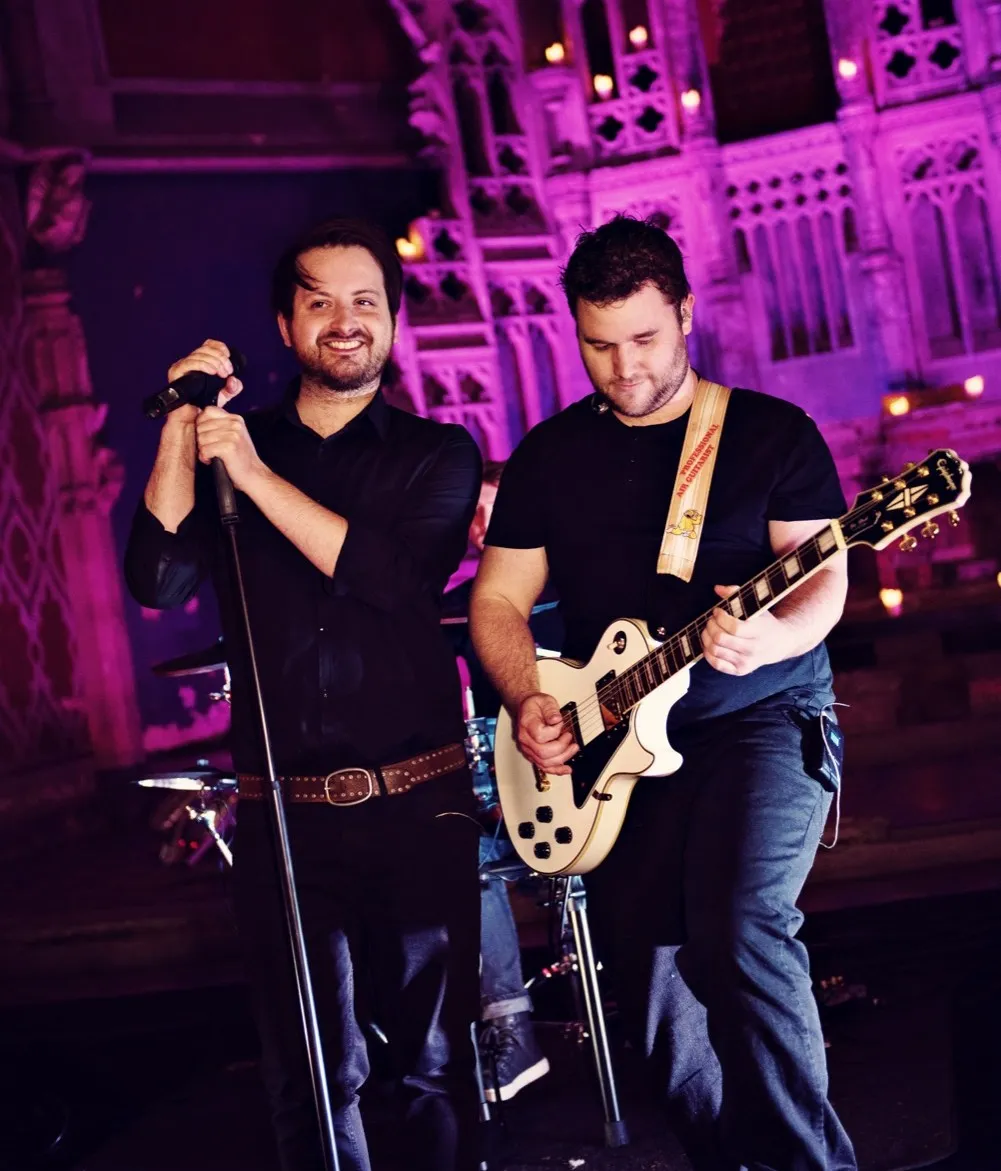 Two male musicians performing on stage, one singing into a microphone and the other playing a white electric guitar, with purple-lit ornate background.