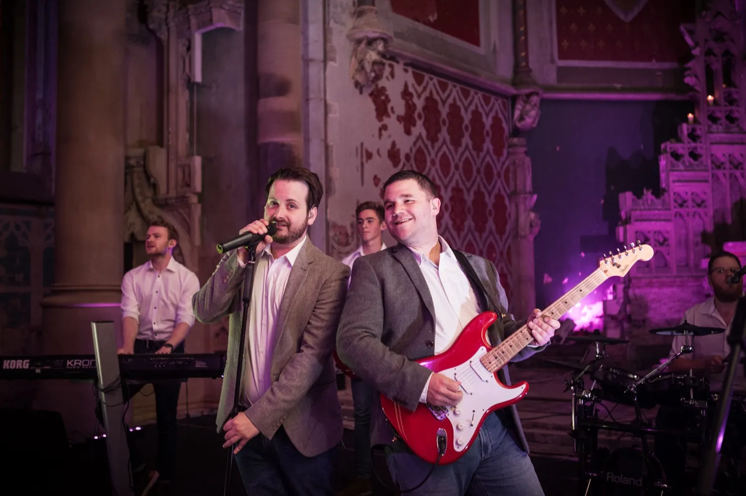 Band performing indoors with a singer holding a microphone and a guitarist playing a red electric guitar.
