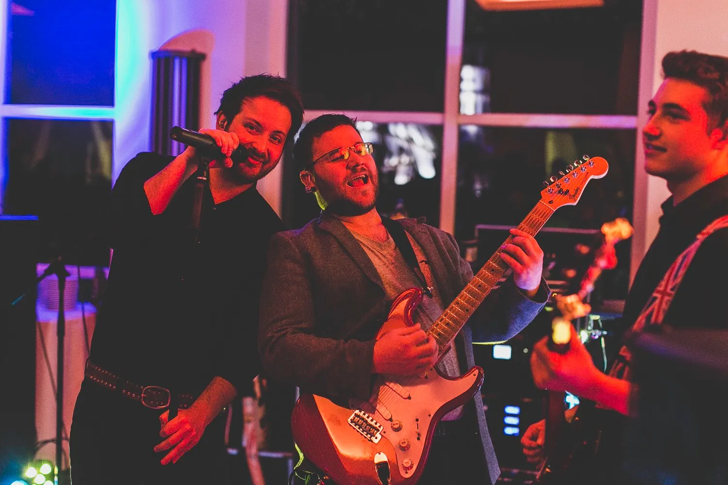 Three men performing live music, one singing into a microphone and two playing electric guitars.