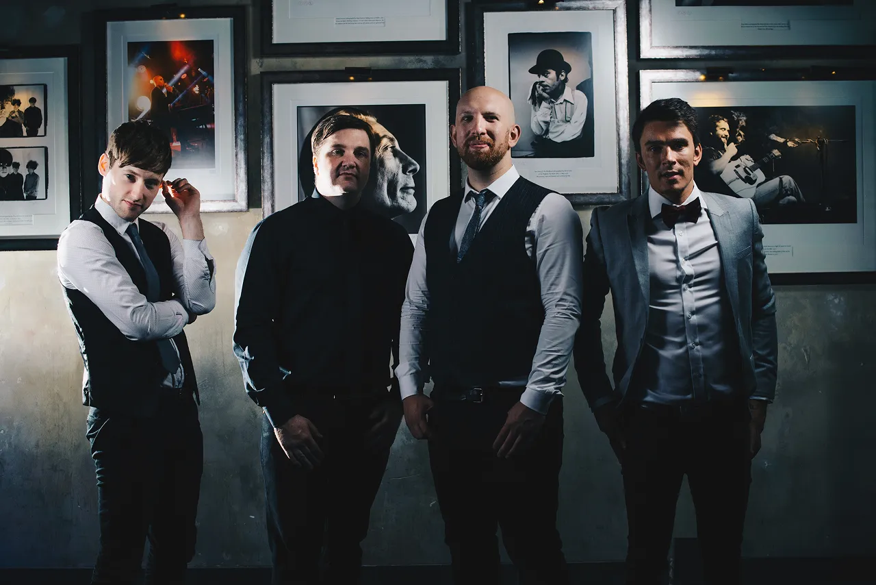 Four men in formal attire standing in front of a wall with framed black and white photographs.