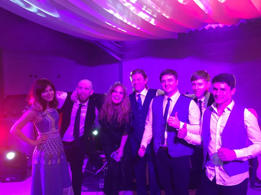 Group of seven people smiling and posing under pink and purple lighting at an indoor event.