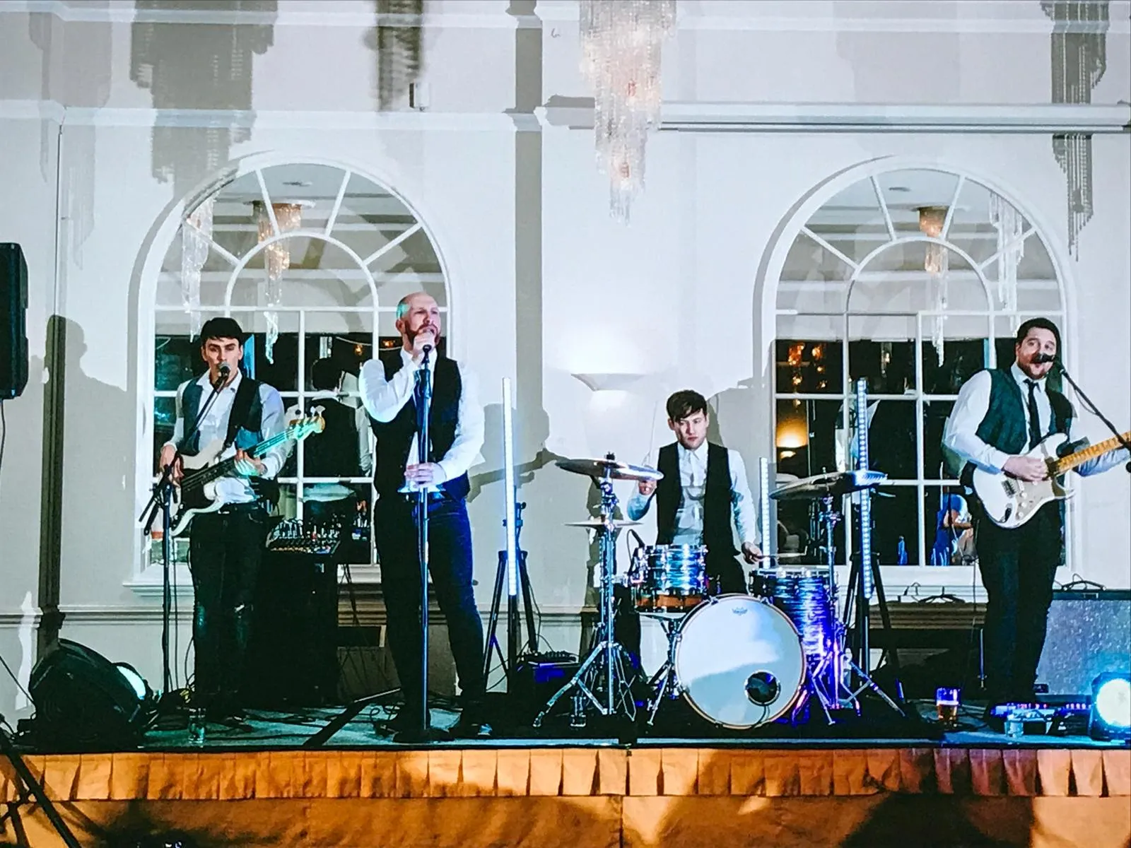 Four-piece band performing on stage with two guitarists, a drummer, and a lead singer in formal vests and white shirts.