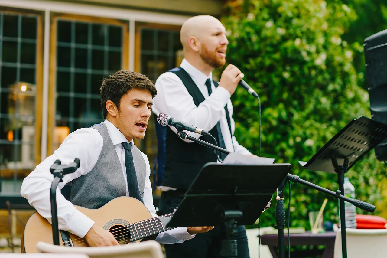 Two men in white shirts and vests performing music outdoors, one playing guitar and singing, the other singing into a microphone.