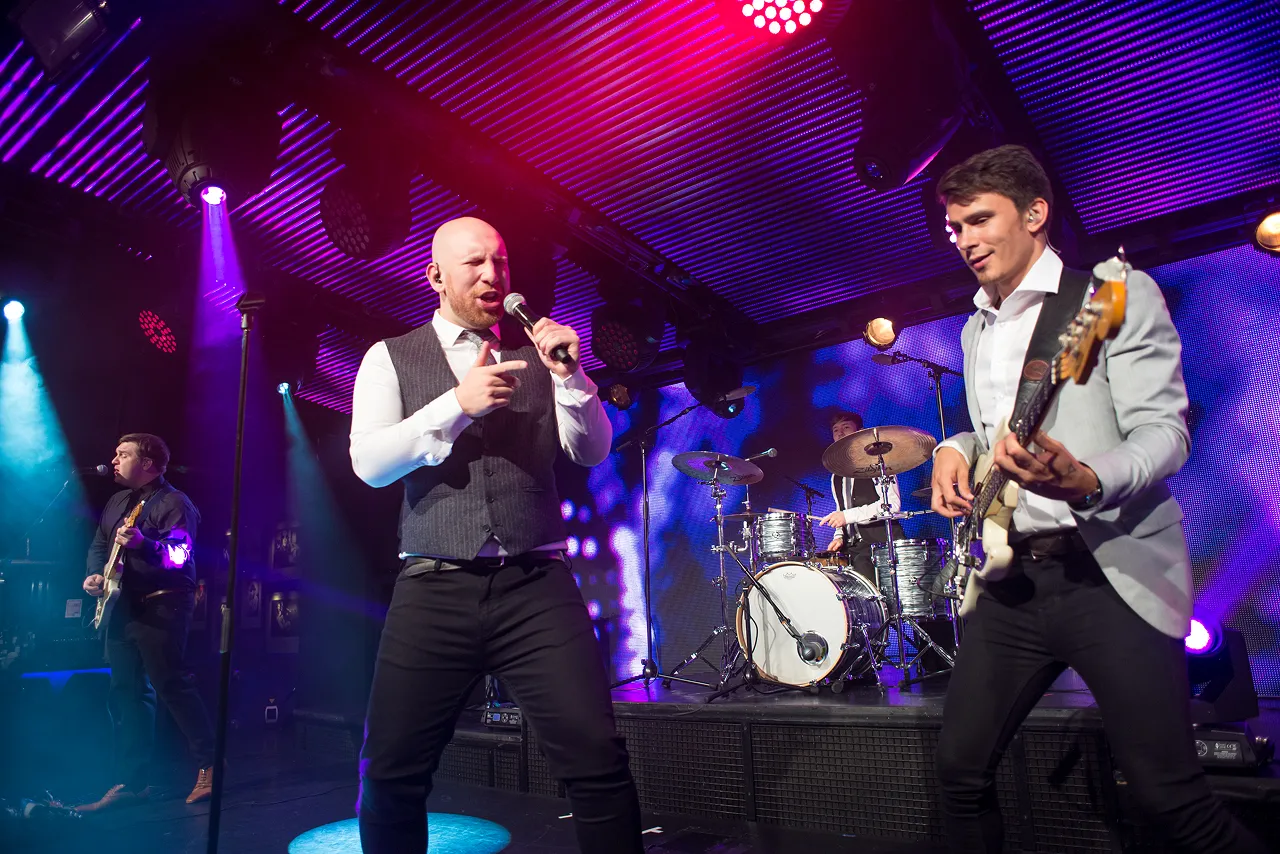 Four-piece band performing on stage with colorful purple and blue lighting, featuring a singer holding a microphone, a guitarist, a bassist, and a drummer.