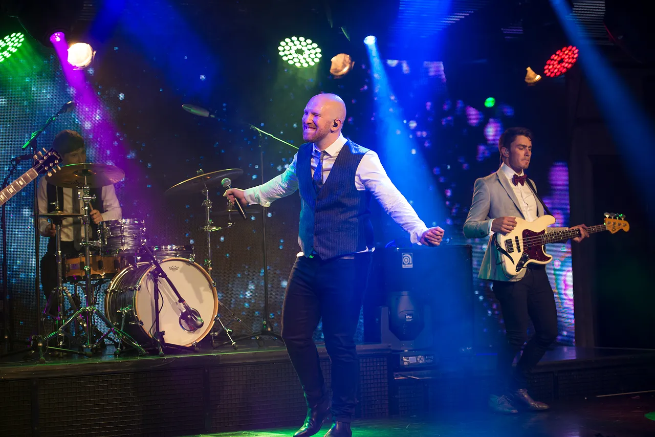 Band performing on stage with a singer holding a microphone, a drummer, and a bassist under colorful stage lights.