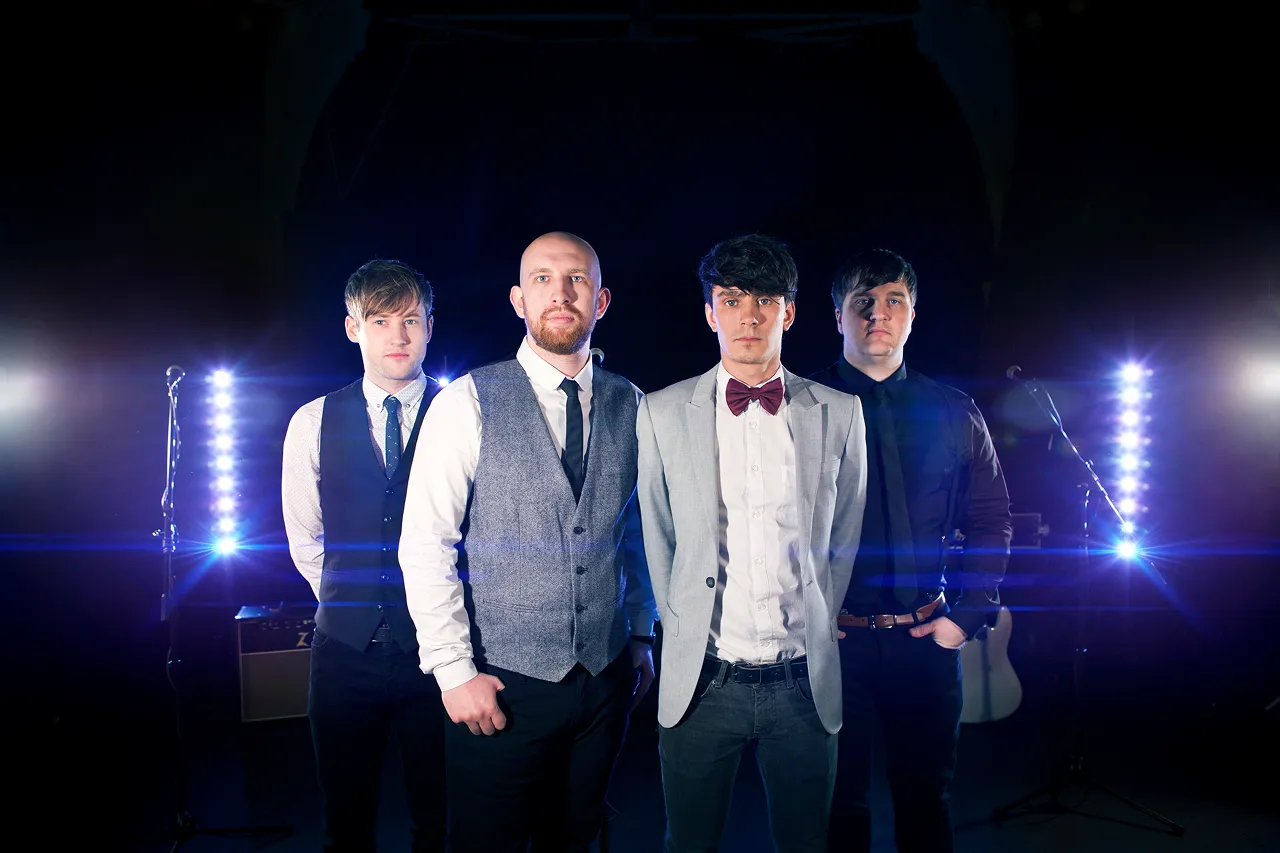 Four men standing in a row in front of stage lights with musical equipment in the background.