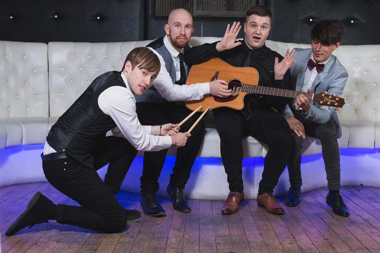 Four men in formal attire posing with a guitar and drumsticks on a white cushioned bench with blue lighting underneath.