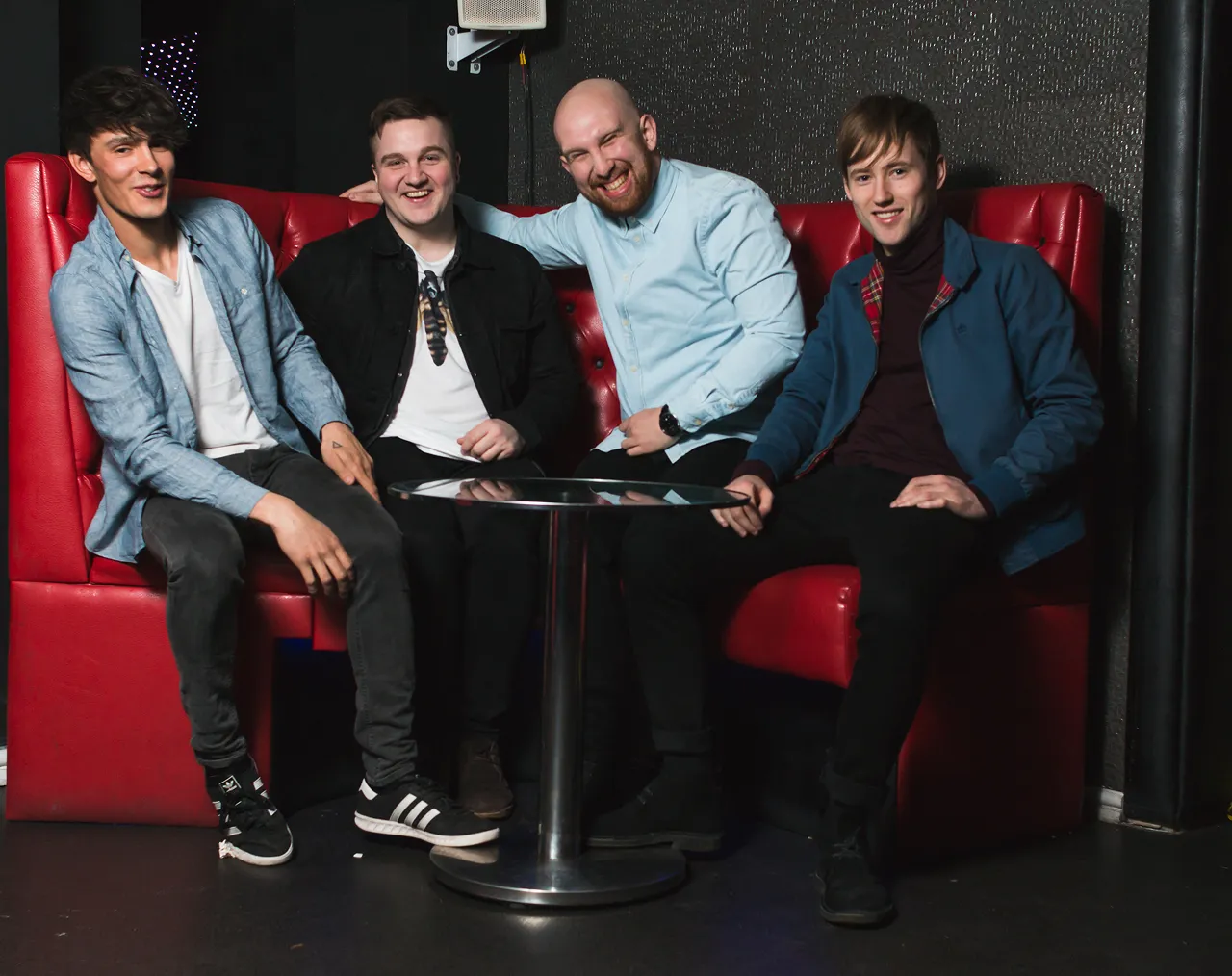 Four young men sitting on a red booth sofa around a round metal table, smiling and posing for a photo.