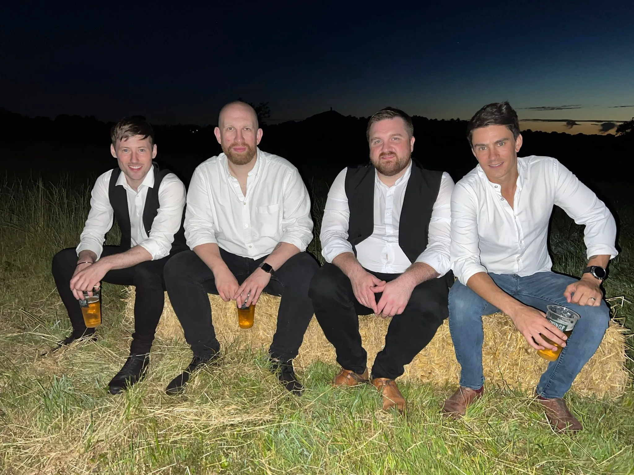 Four men dressed in semi-formal white shirts and black or blue pants sitting on straw bales outdoors at night, each holding a pint of beer.