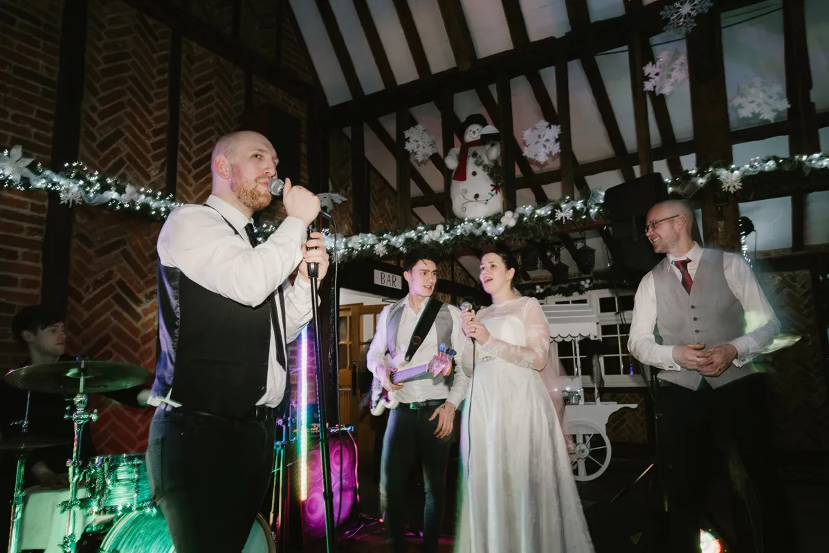 Wedding band performing indoors with a female singer in a white wedding dress and musicians playing instruments under festive decorations including a snowman and garlands.