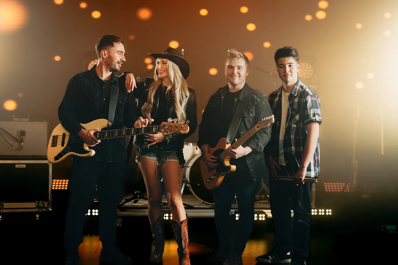 Four musicians standing on a stage with instruments, including two guitars and drumsticks, smiling in warm stage lighting.