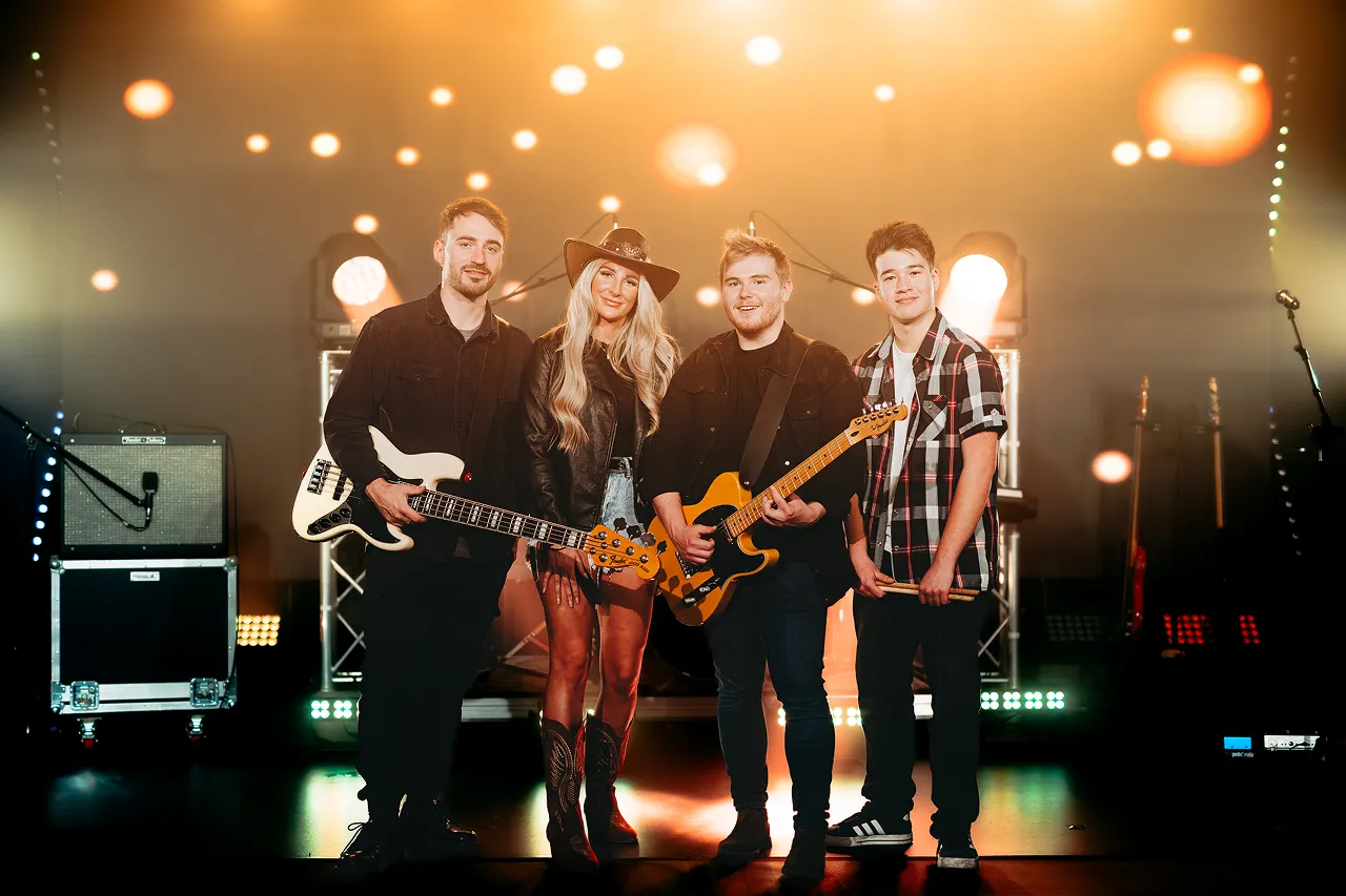 Four young musicians standing on stage with guitars and drumsticks under warm stage lights.