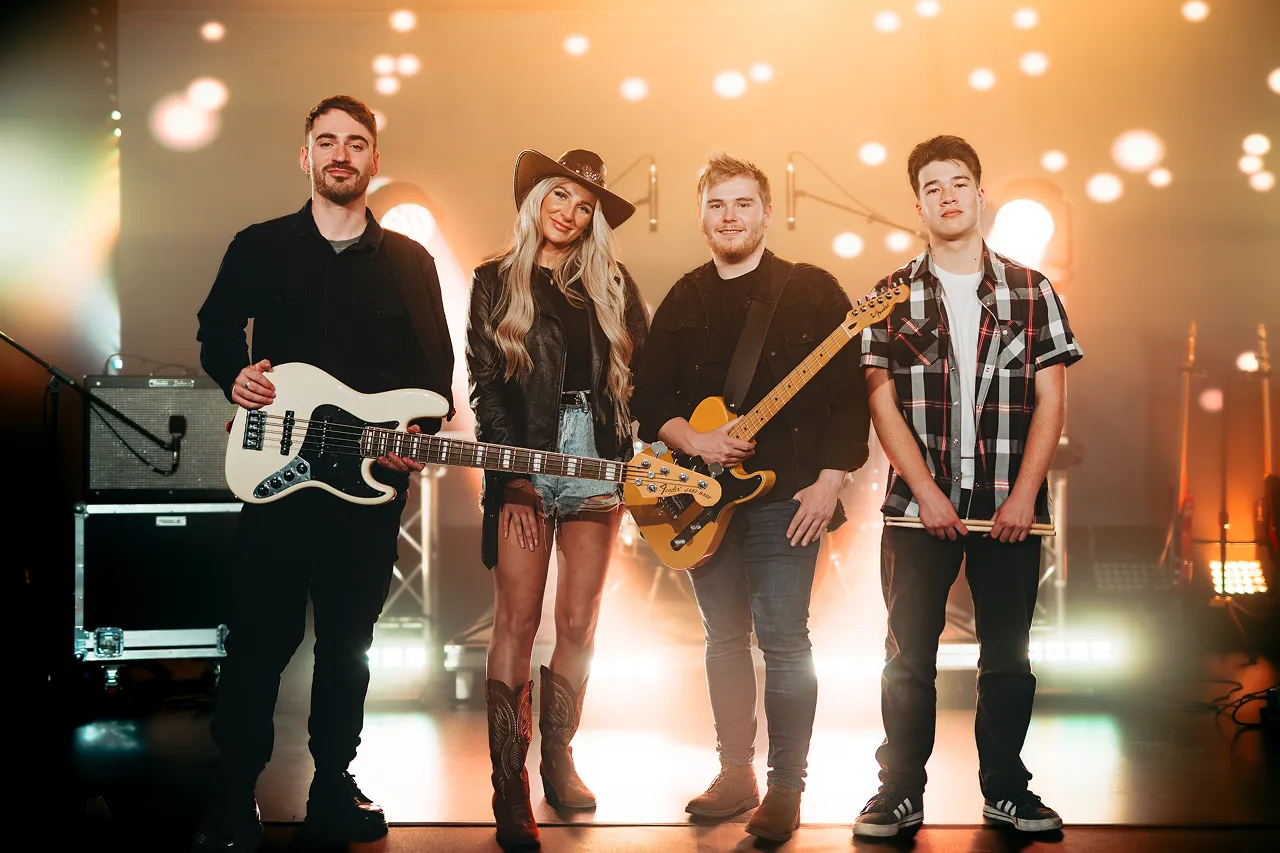 Four musicians standing on a stage with instruments and warm bokeh lights in the background.