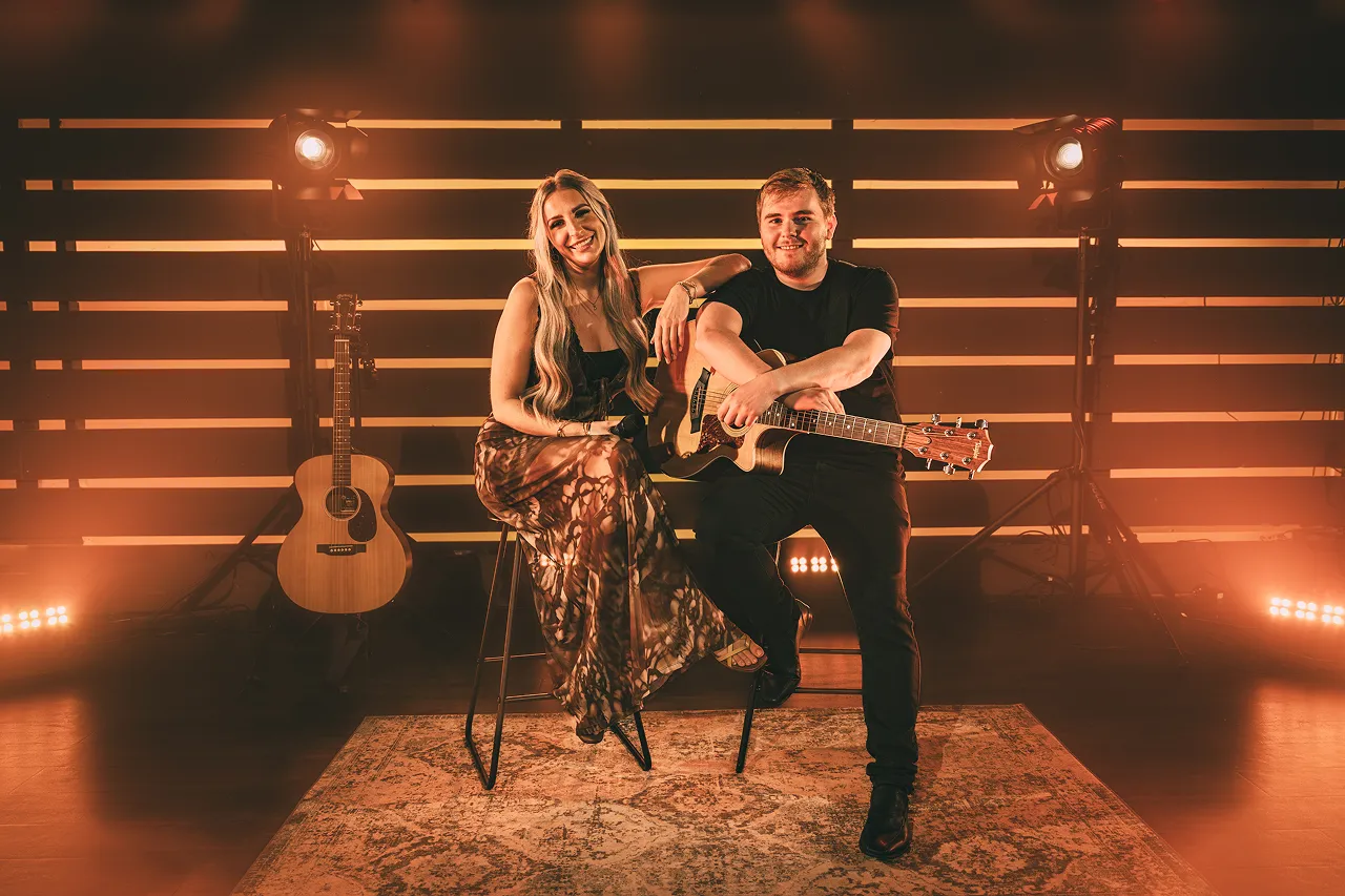 A man and a woman sitting on stools; the man holds an acoustic guitar while the woman smiles with her arm resting on his shoulder, two guitars and stage lights in the background.