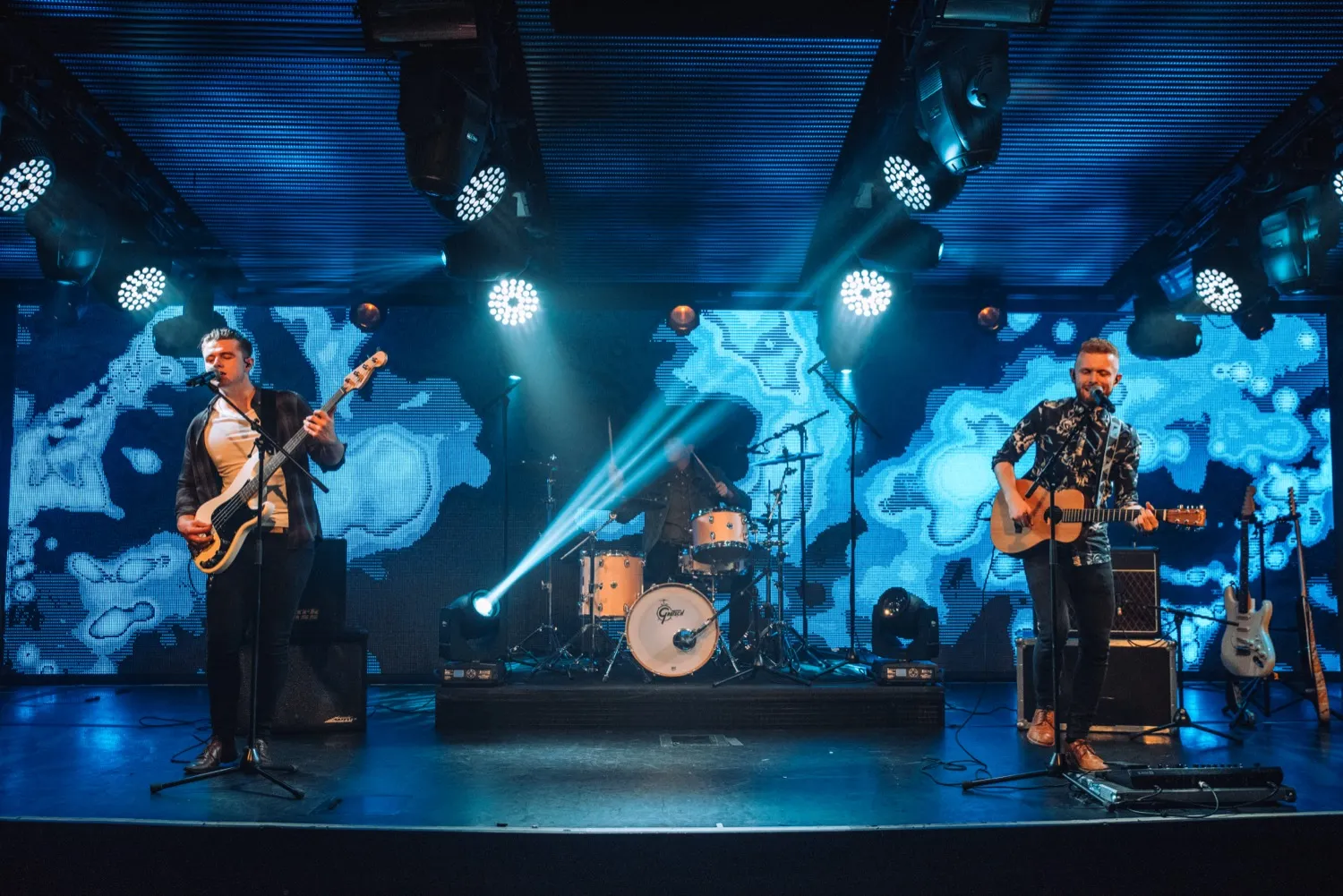 Three-piece band performing on stage with electric bass, drum set, and acoustic guitar under blue stage lights.