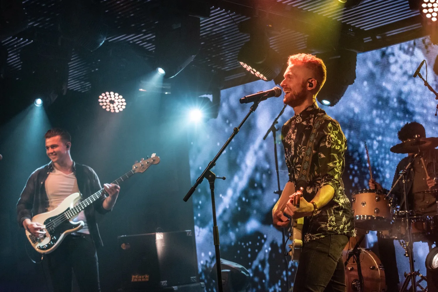 Band performing live on stage with a guitarist singing into a microphone and a bassist smiling.