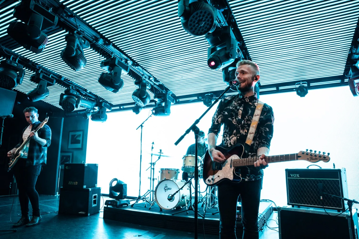 Three-member band performing on stage with electric guitars and drum set under blue stage lights.