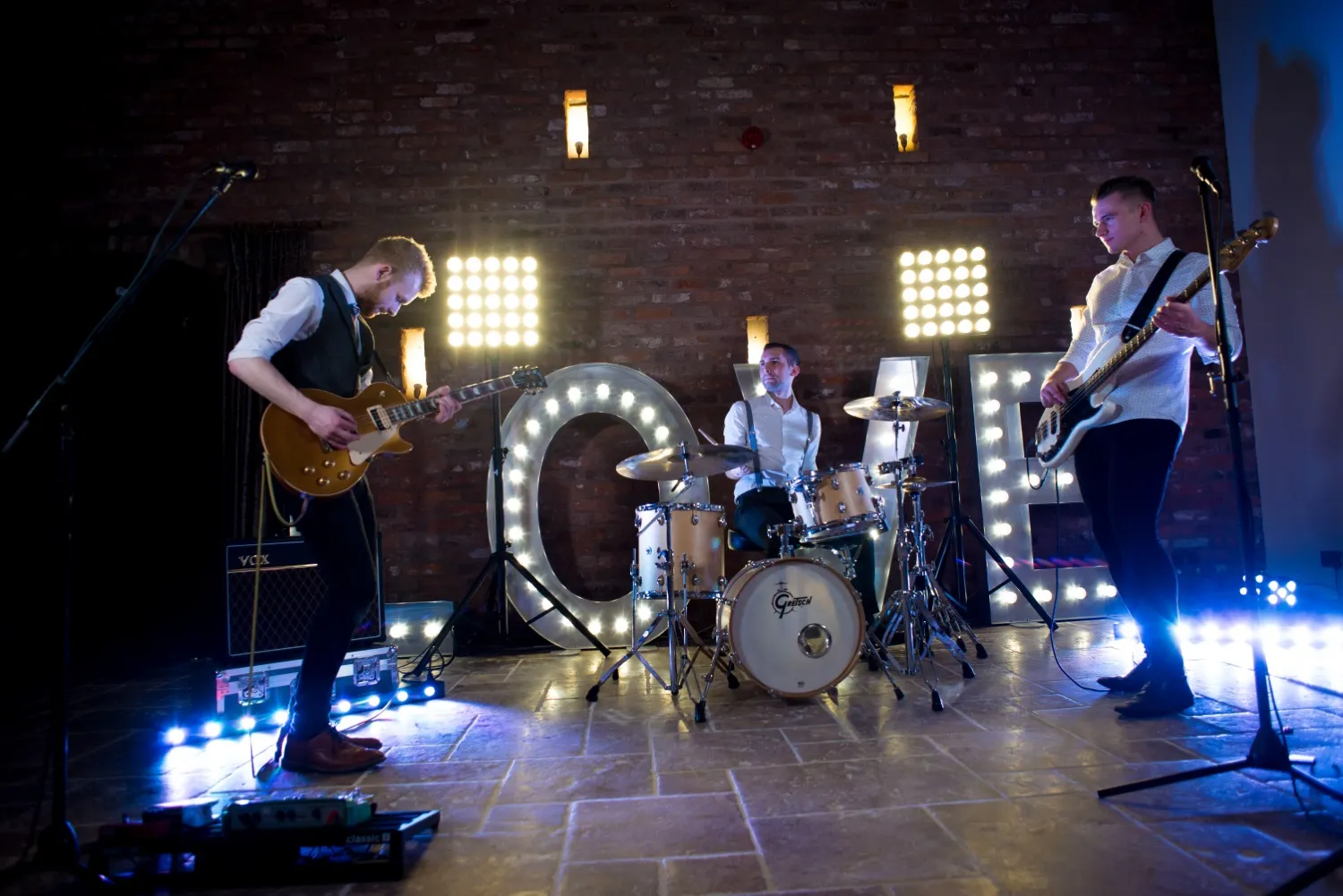 Three-member band performing indoors with a guitarist, drummer, and bassist, illuminated letters spelling LOVE in the background.