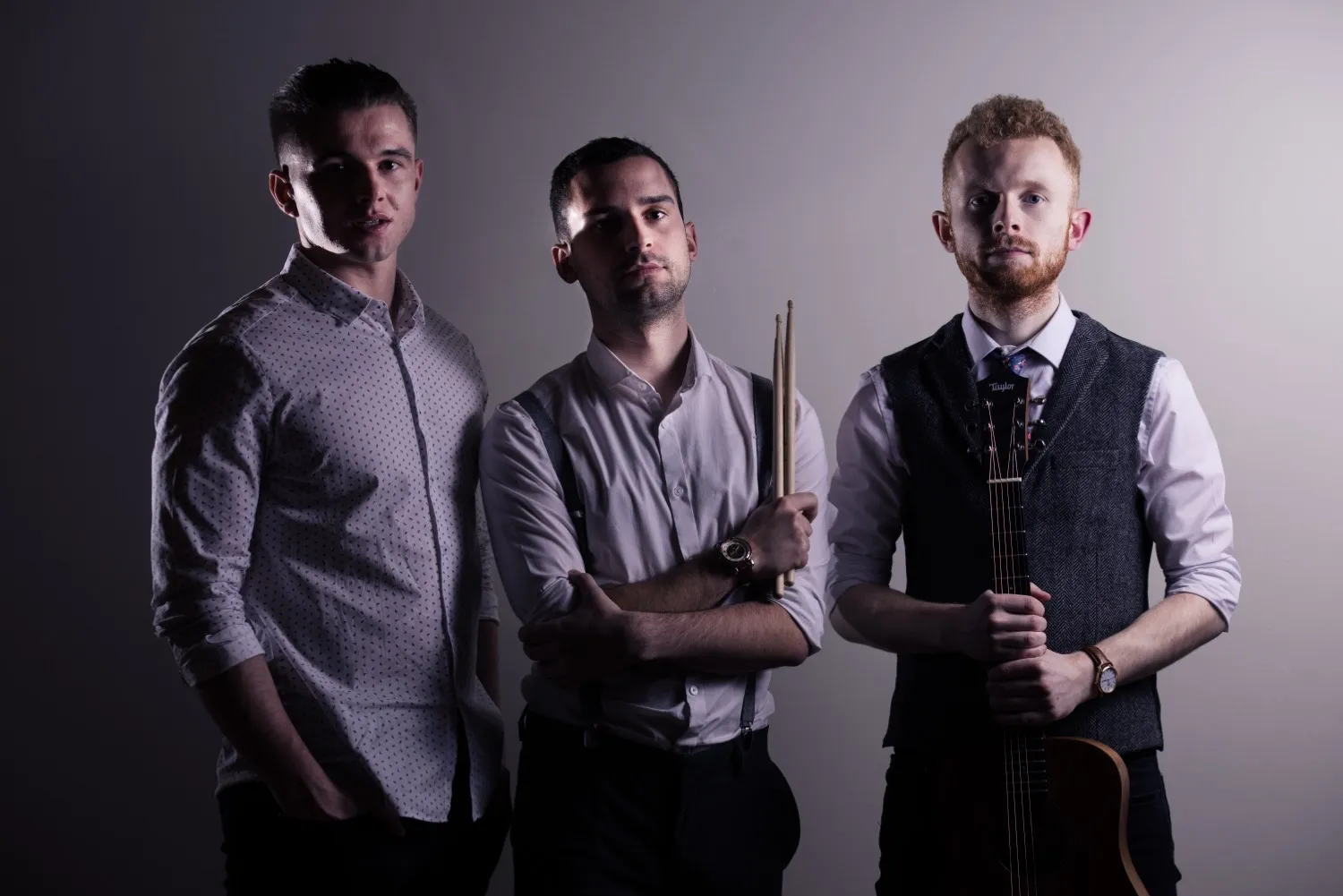 Three men posing against a neutral background, one holding drumsticks and another holding an acoustic guitar.