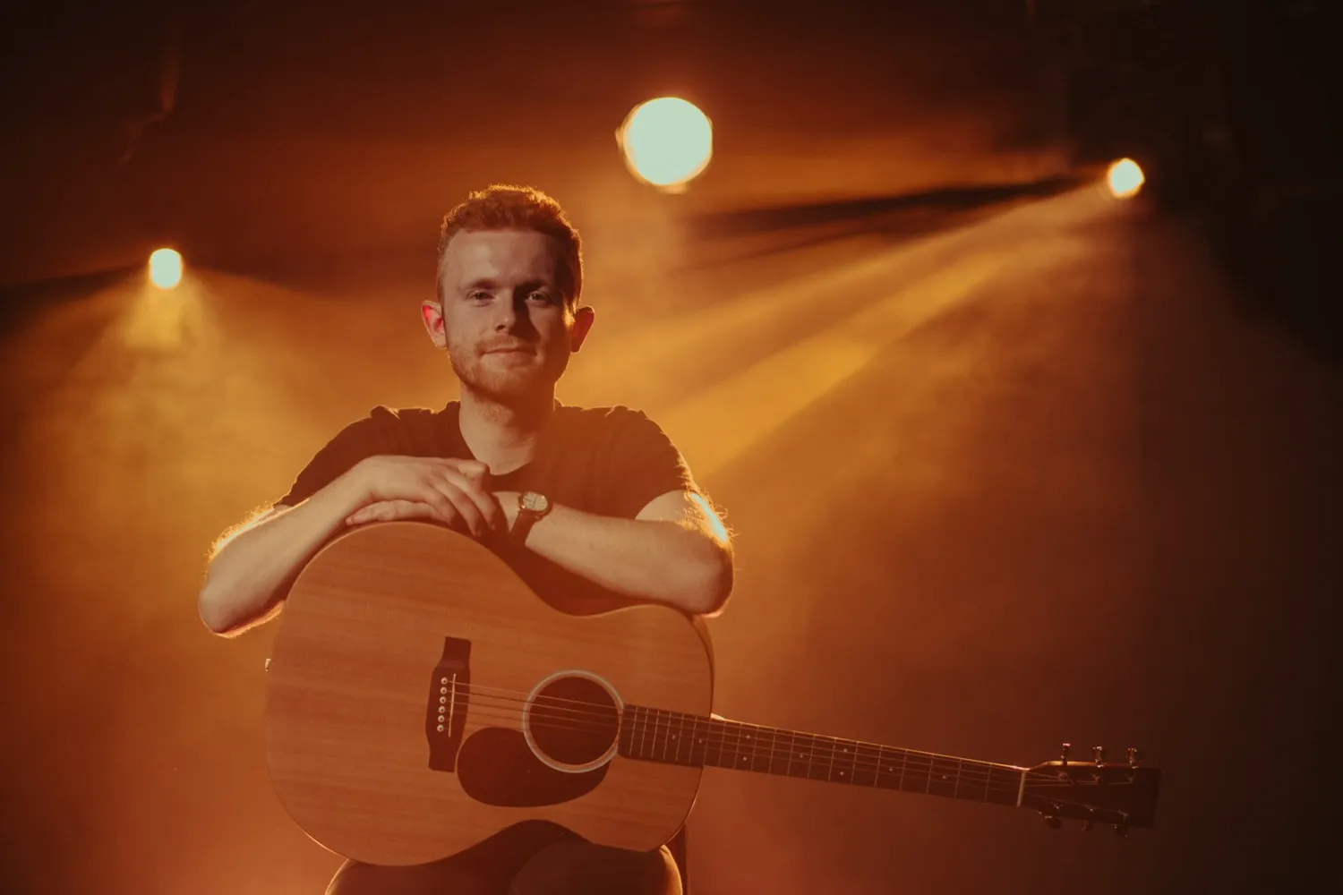 Man sitting with an acoustic guitar, illuminated by warm stage lights in a smoky atmosphere.
