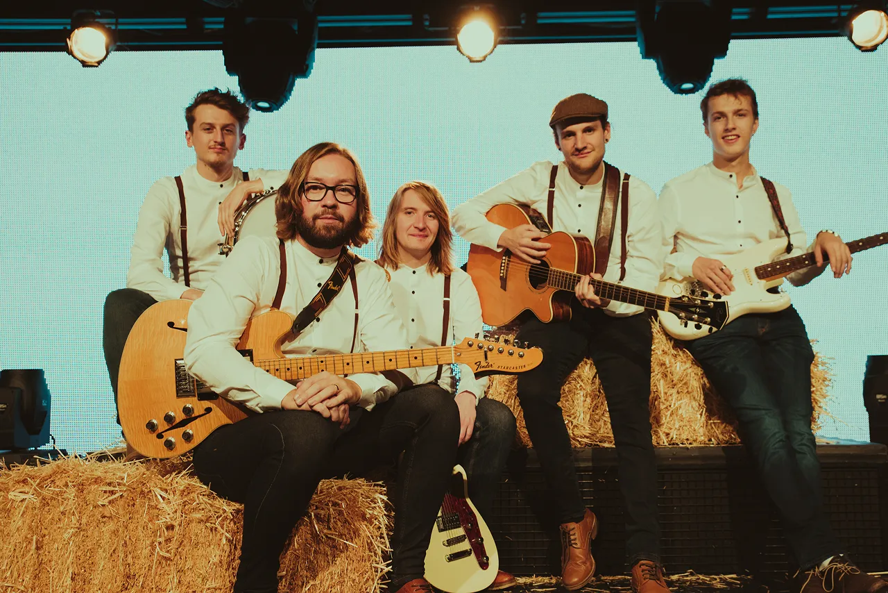 Five male musicians in white shirts with suspenders sitting on straw bales, holding guitars and a drum on stage.