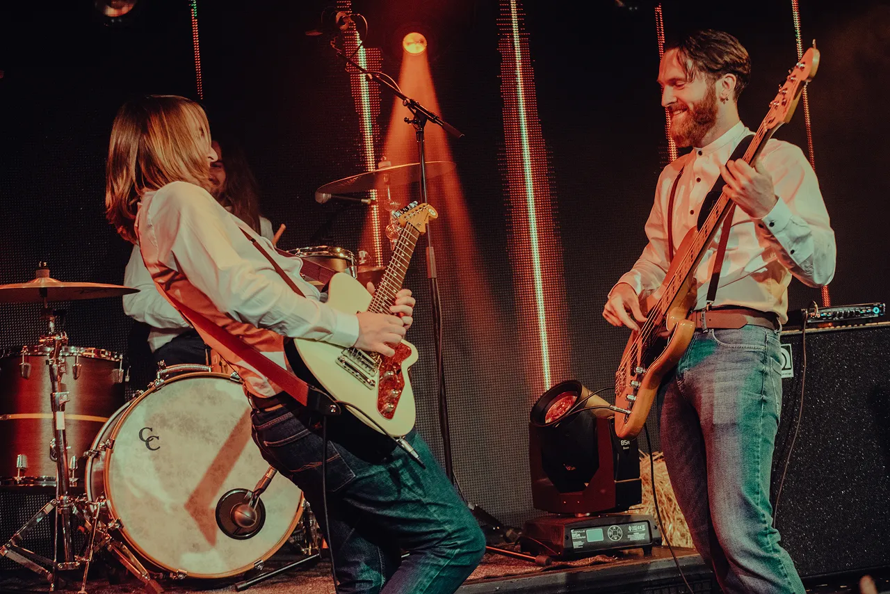 Two male musicians playing electric guitar and bass on stage with drums in the background under warm lighting.