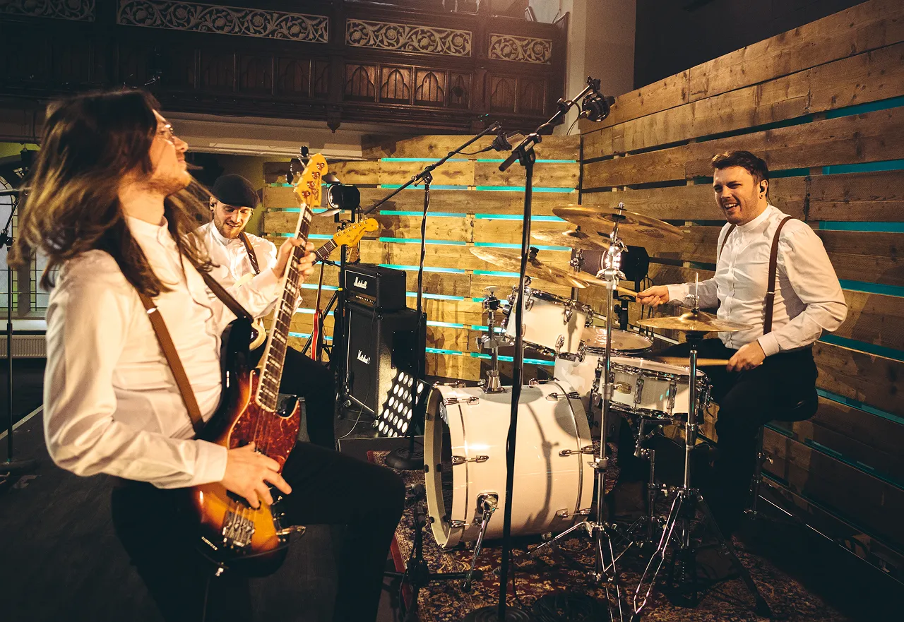 Three musicians performing in a studio with wooden paneling, one playing drums and two playing guitars.
