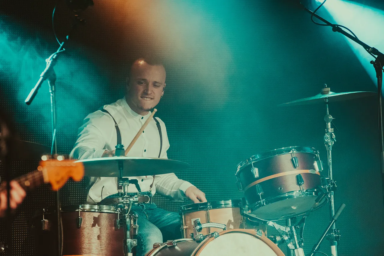 A drummer wearing a white shirt and suspenders playing a drum set on stage under blue and orange stage lights.