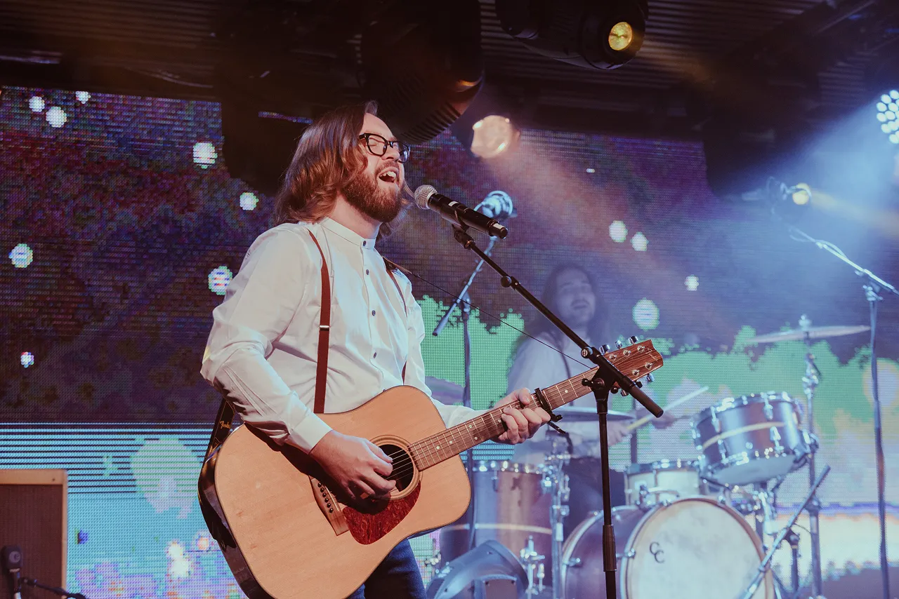 Male musician with glasses and beard singing and playing acoustic guitar on stage, drummer blurred in background.