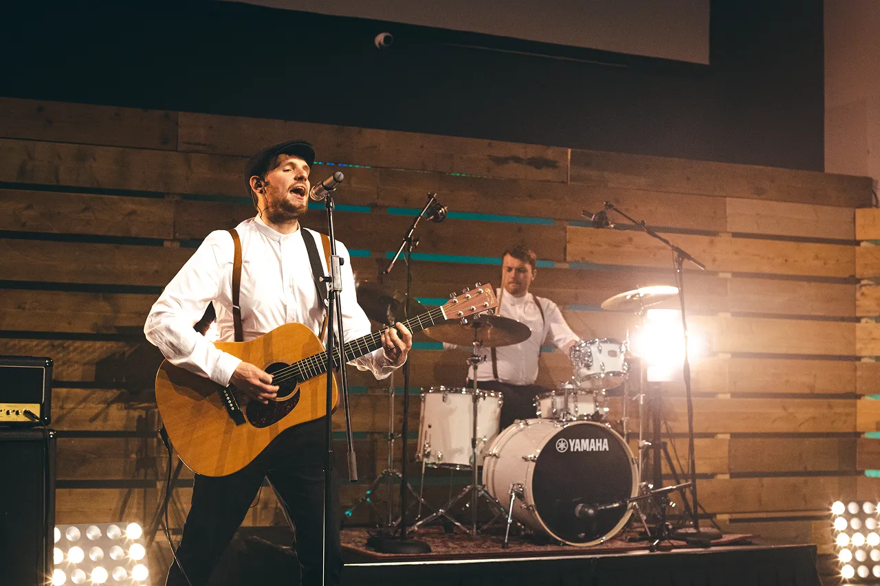 Male musician singing and playing acoustic guitar with drummer in background on stage with wooden panel backdrop.